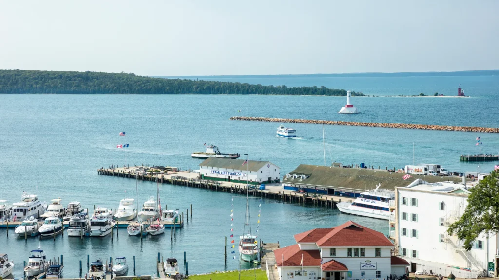 A harbor in Mackinac Island with boats lined up side by side and a small light house in the distance. The water is a dark teal and small aged buildings peak out in the corner of the photo.
