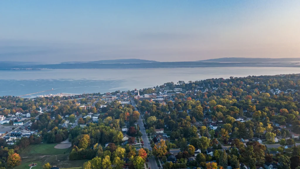 An aerial of Petoskey, MI showing the Great Lake, Lake Michigan, and the residential areas surround the lake. The sky is dusky in an unsaturated blue that transitions into a pale peach color.