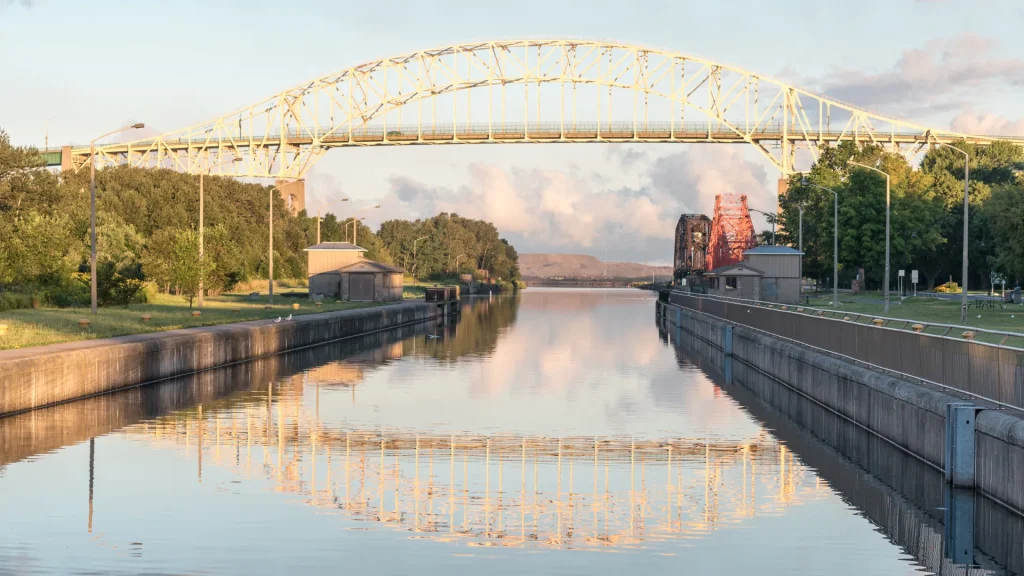 The Sault Ste. Marie bridge crossing the canal in Sault Ste. Marie. The canal is still with a glass-like imagery as the bridge is reflected perfectly in the water. On each side are trees and green grass as it opens out into the water. The sky is a pale robin's egg blue with fluffy pink tinted clouds.