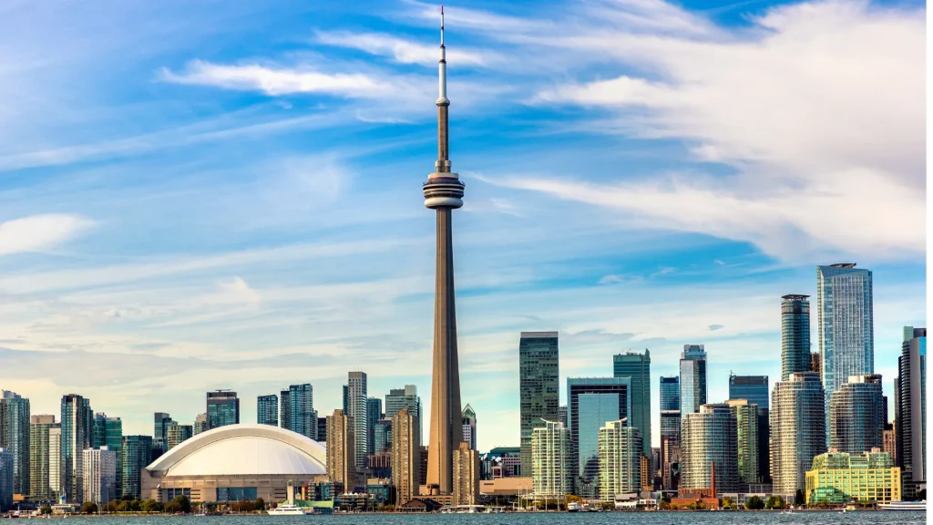 The city skyline of Toronto, CA with a tall, slender, tower in the middle calling focus. The sky is bright blue with wispy clouds. The water of the lake is set at the bottom of the photo in the foreground.