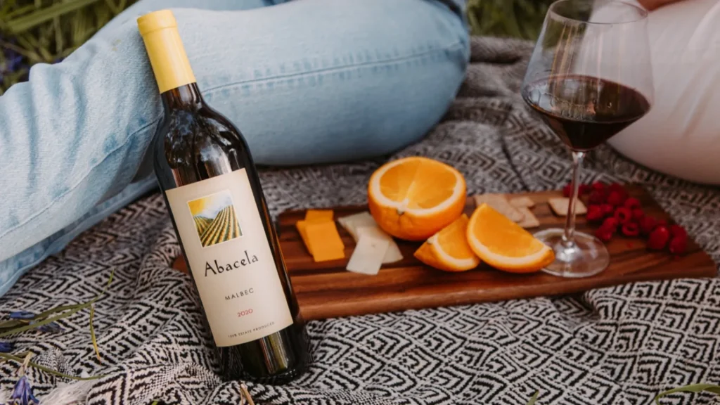A bottle of Abacela wine rests against a person's knee as it sits on a picninc blanket with a charcuteie board of orange, a variety of cheses, raspberrys, crackers and a glass of red wine.