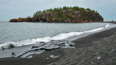 Black Beach in Minnesota. The day is overcast as the water washes up onto the black beach. An island sits in the distance.