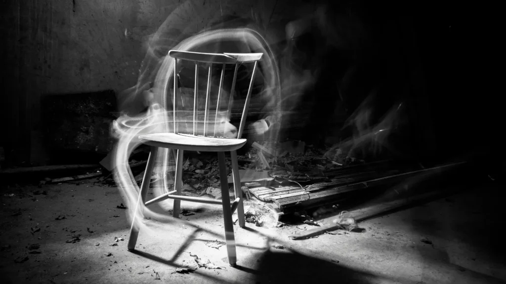A black and white photo of an old wooden chair in a dilapidated home as a swirl of smoke or energy move around it.