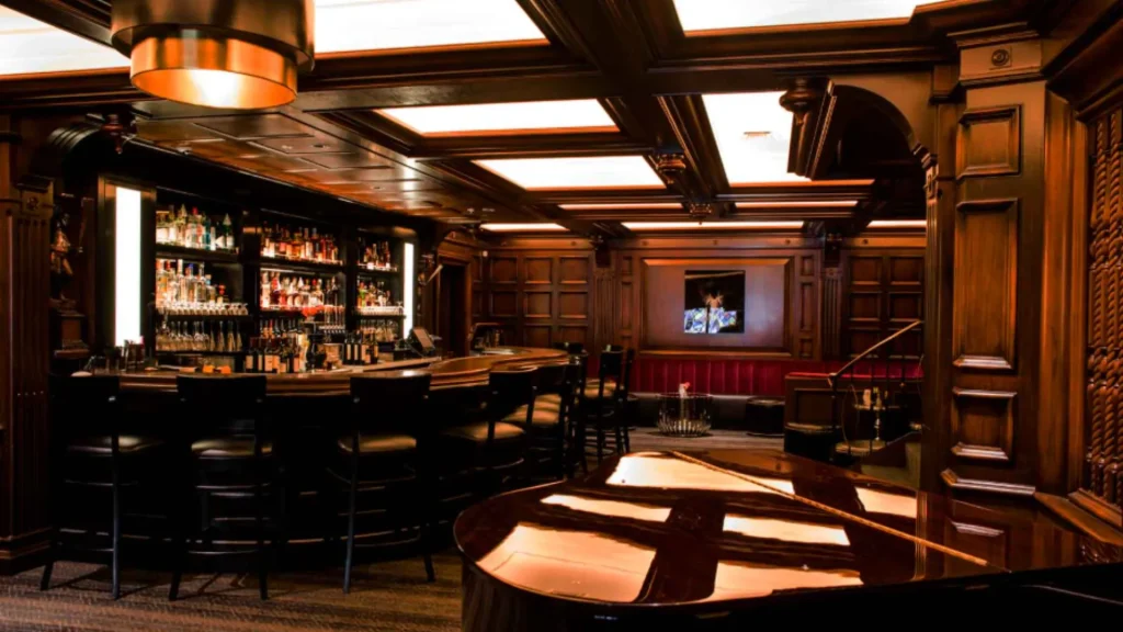 The bar in The Lexington is classically styled with wooden paneling on the ceiling and walls. A piano sits in the corner of the photo as well. 