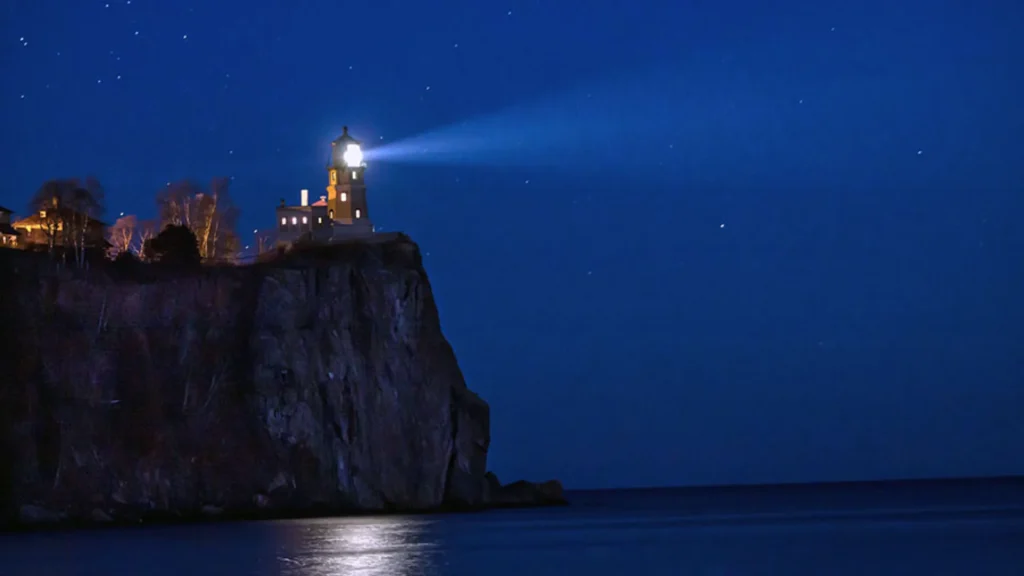 The Split Rock Lighthouse State Park at night. The lighthouse is light up brightly against the deep cobalt blue sky and water. 