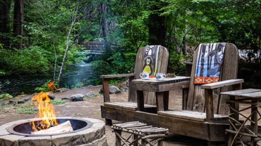 A wooden chair bench, separated by a side table all built into one piece sits in front of a stone outdoor fireplace and a river.