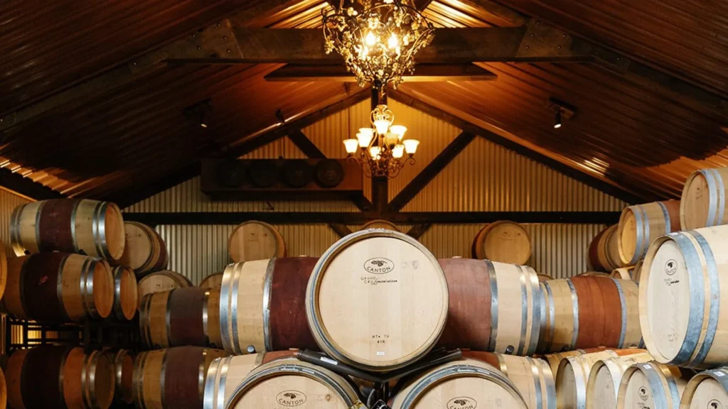 Barrels stacked upon each other in a room built with metal sheets and wooden beams. A chandeleir hangs down from the center.