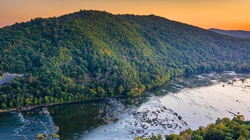 The sunset view from an outlook on the Weverton Cliffs hike. The sky is a citrus orange, which contrasts with the deep green and blue tones of the forested range.