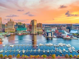An aerial photograph of Baltimore, MD at sunset. The sky is orange, yellow, and pink with clouds dotting across it.