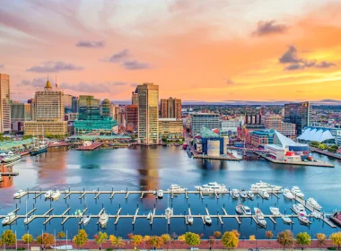 An aerial photograph of Baltimore, MD at sunset. The sky is orange, yellow, and pink with clouds dotting across it.