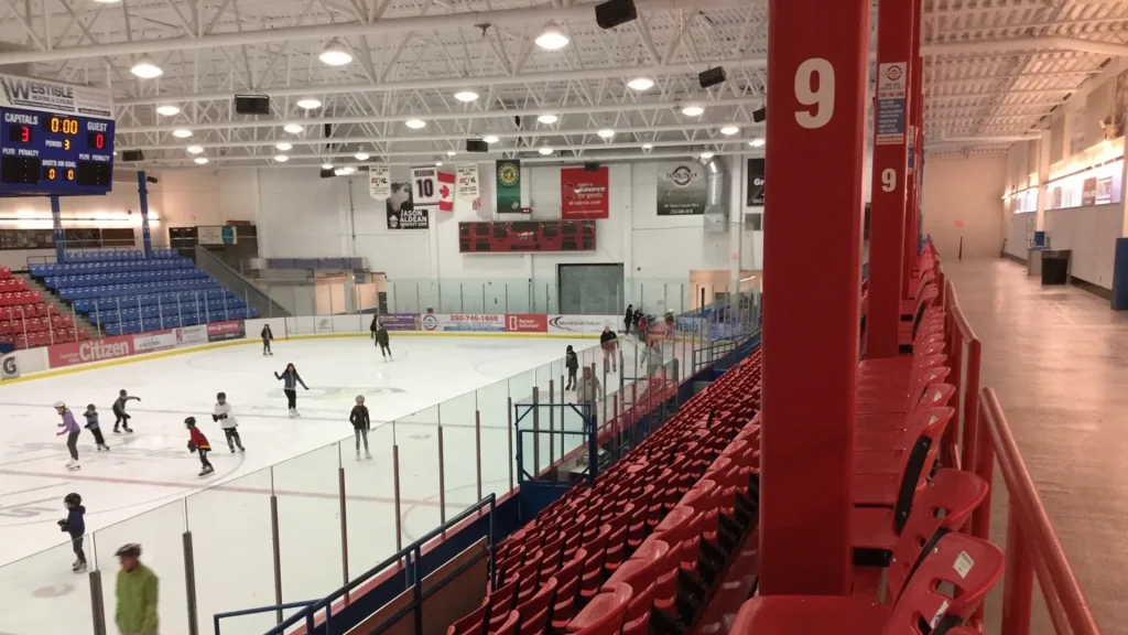 The Cowichan Community Centre in B.C., where the Duncan Stick is attached. Here we see the interior of the Community Centre and an ice rink with people skating upon it. It's surrounded by red bleacher style seating. 