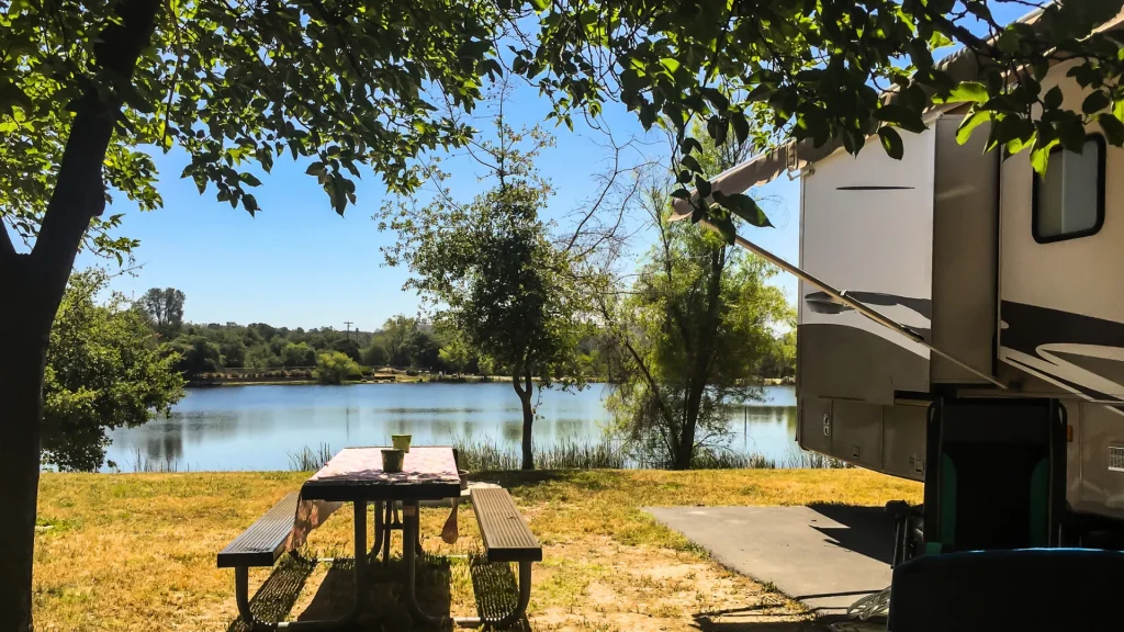 An RV set up beside a picnic table that looks out to a lake on a sunny day.