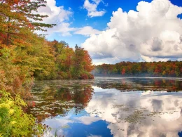 Tobyhanna State Park in the fall. The photo depicts a lake surrounded by fall colored foliage on a sunny yet cloudy fall day.
