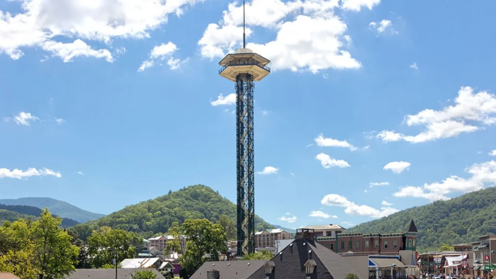 A ground view image of the Gatlinburg Space Needle during the day as it towers over the city into the bright blue sky, dotted with clouds.