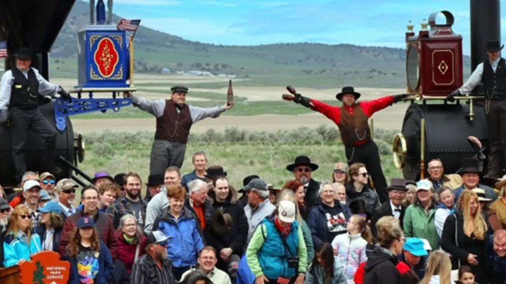 Two people hang off the front of two separate locomotives facing each other as a crowd of people stand in front of them. 