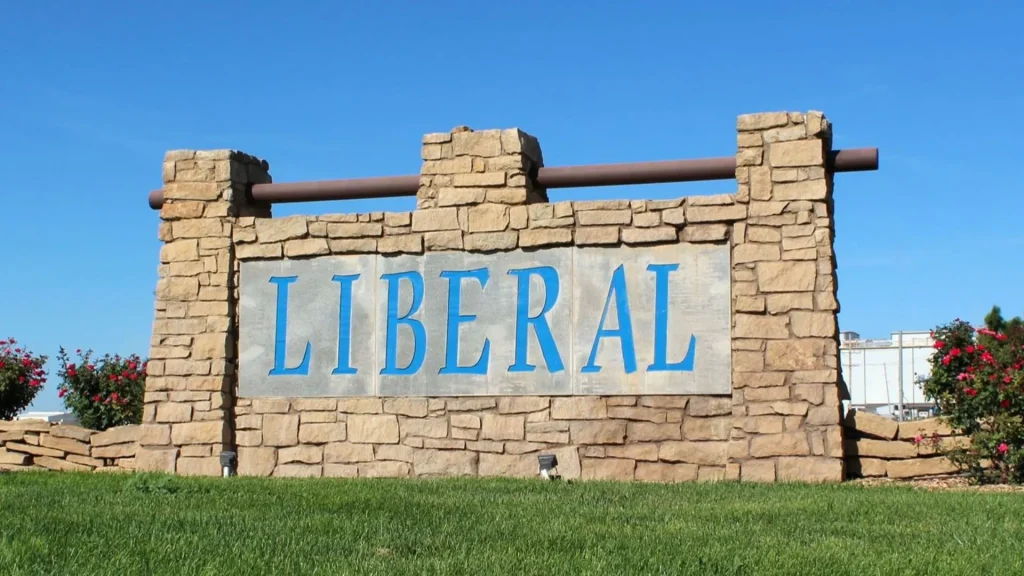 Liberal City's sign is in bricks with LIBERAL in blue letters. The day is sunny, and the sky is clear and bright blue as the sign protrudes from green grass.