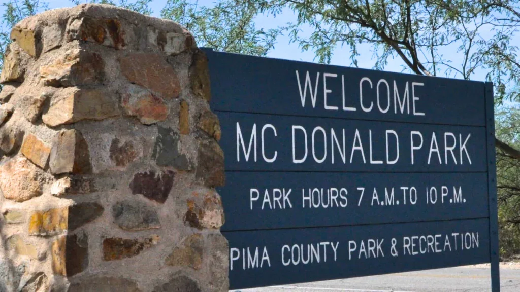 The park sign for McDonald Par listing it's hours in white text against the blue painted wooden sign. It's held up by a stone pillar and set with concrete at the bottom. The sign reads: WELCOME MC DONALD PARKPARK HOURS 7 A.M. TO 10 P.M.PIMA COUNTY PARKS & RECREATION". 