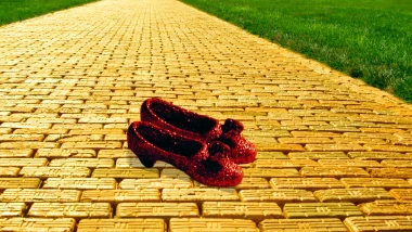 Two red sequin slippers, akin to Dorothy's from Wizard of Oz, sit on a yellow brick road with green grass on each side of it.