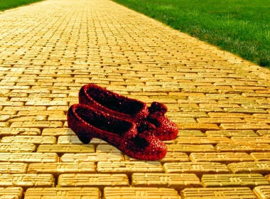 Two red sequin slippers, akin to Dorothy's from Wizard of Oz, sit on a yellow brick road with green grass on each side of it.