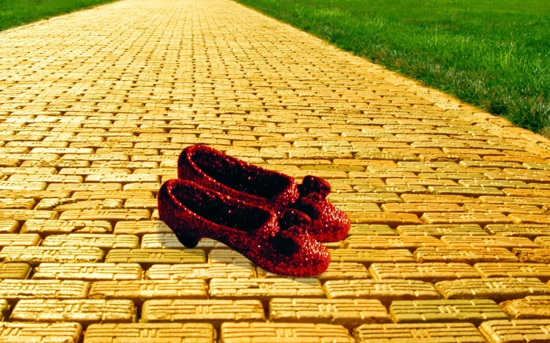 Two red sequin slippers, akin to Dorothy's from Wizard of Oz, sit on a yellow brick road with green grass on each side of it.
