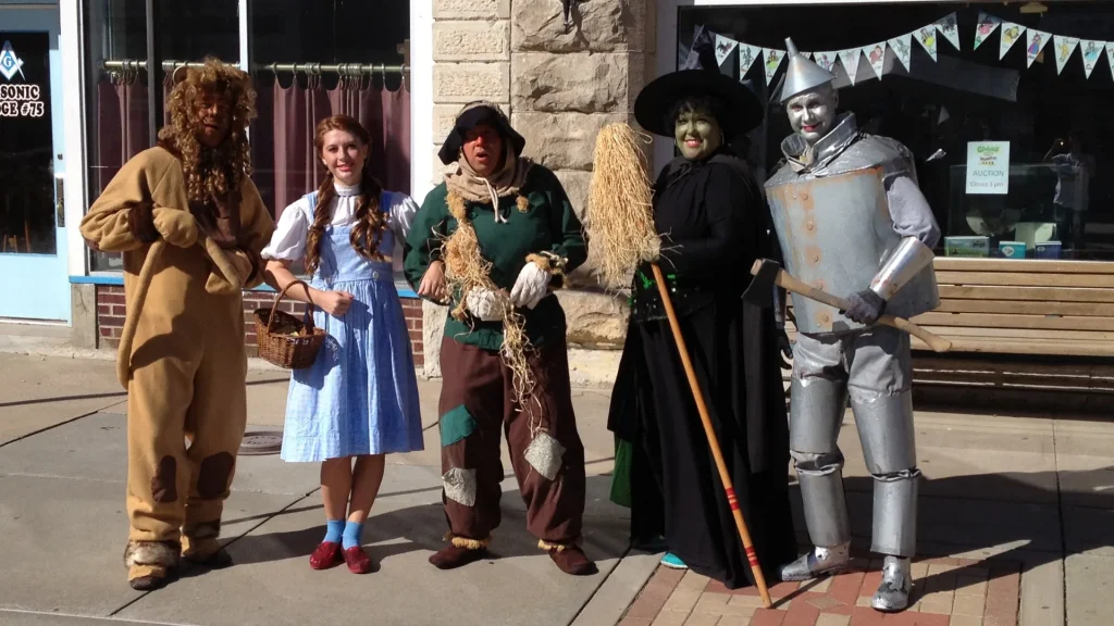 A group of five individuals dressed up in costumes as Wizard of Oz characters: Lion, Dorothy, Scarecrow, Wicked Witch, and Tin Man. They stand together for the photo posing.