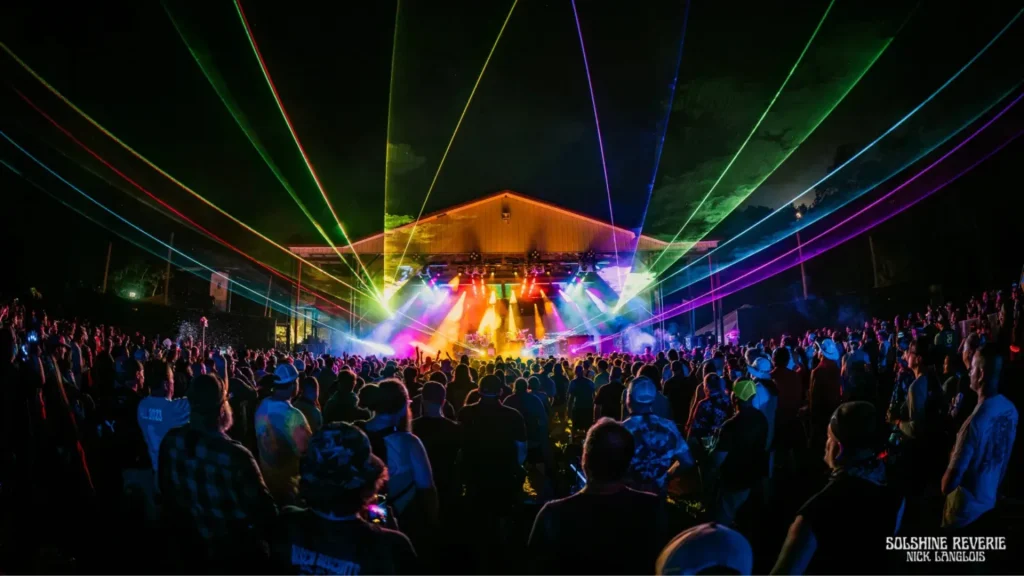 A night photo of an outdoor music festival as a laser light show streams lights in all colors above a crowd of people.