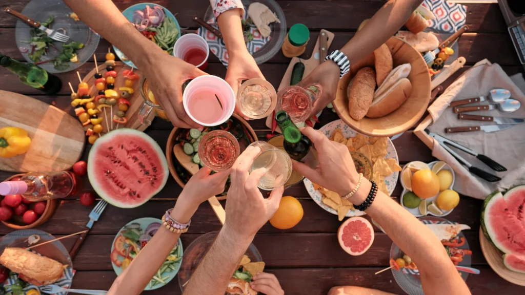 An aerial photo of the arms and hands at a picnic table of people all tipping their drinks towards each other in a cheers over an abundant amount of food.
