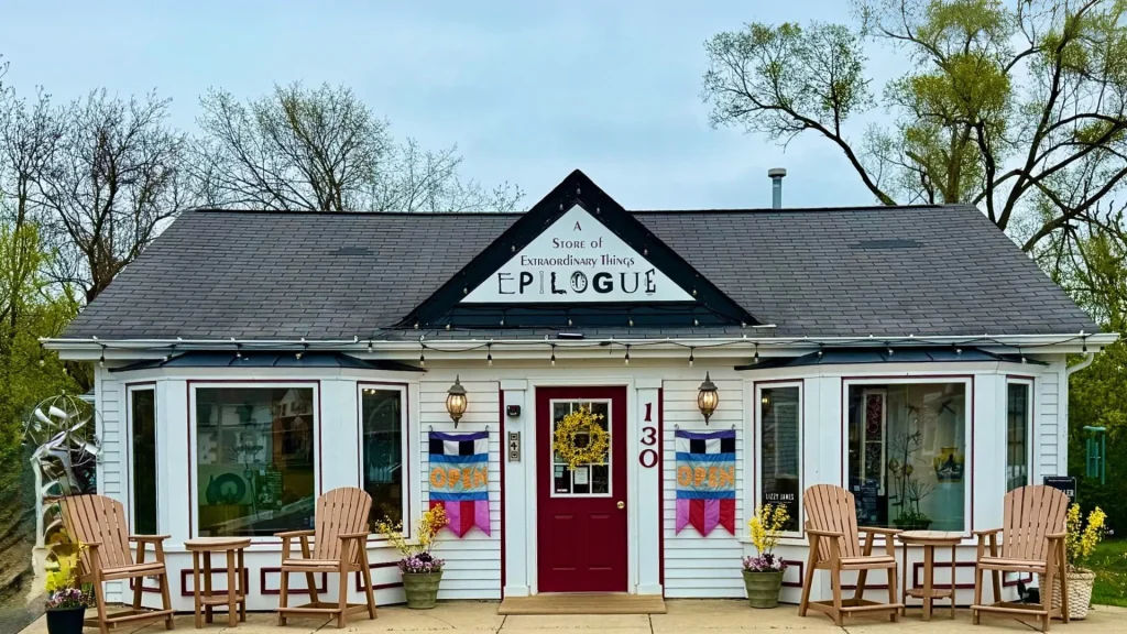 An art store in Long Grove called Epilogue has a quirky sign in the small white building. The front door is a deep red, and sitting out front are wooden chairs and tables. 