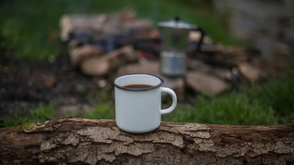 A photo of a cup of coffee sitting on a log with a coffee press in the background, out of focus, near a campfire.