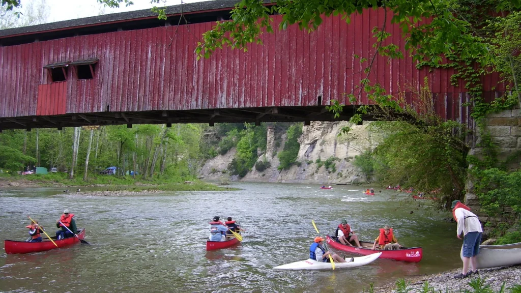 A photo of Clements Canoes Outdoor Center canoes out on a river beneath a red bridge. They launch and slowly drift down the river in red and white canoes.