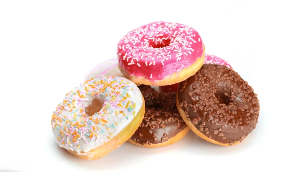 A stack of donuts in chocolate, vanilla, and strawberry frosting with sprinkles, sit atop each other against a white backdrop.