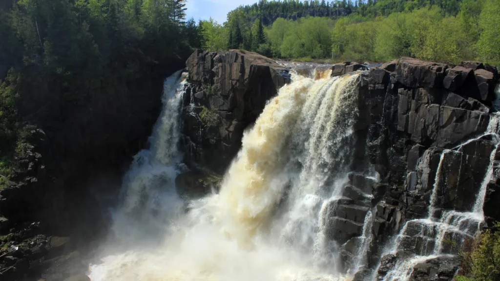A photo of the tallest waterfall in Minnesota, the High Falls of Pigeon River. The rockface is dark, almost black, as the water rushes powerfully over the top and crashes into a white mist at the bottom.