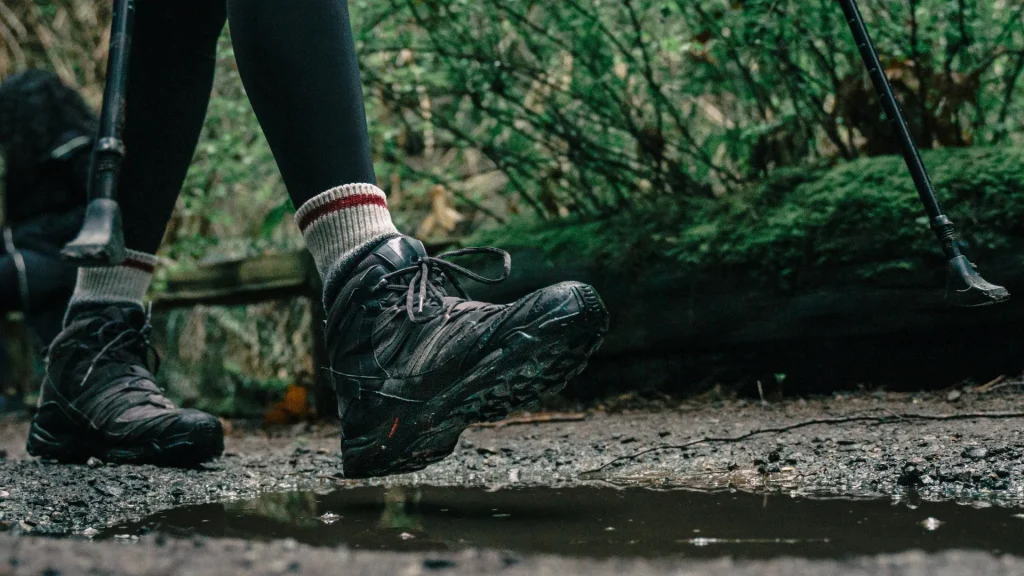 A close up photo of a person's hiking boots as they walk a trail in the woods, about to step into a muddy puddle.