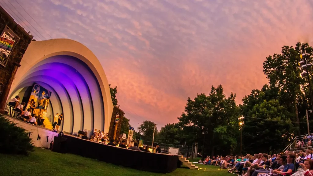 An amphitheater in Lindsborg, KS lit up with purple lights as people gather around it in the grass and on bleacher-style seating.