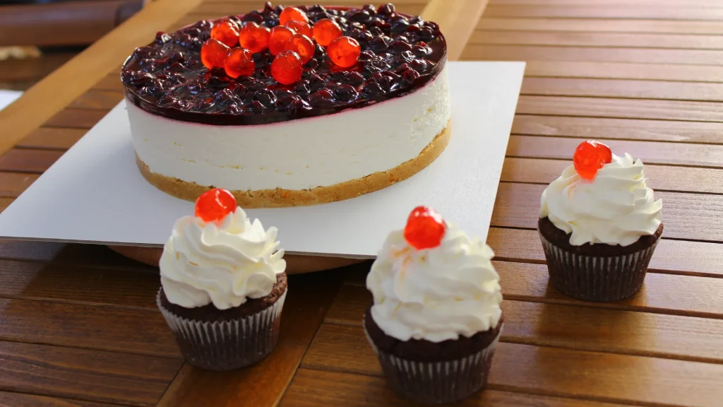 A cheesecake and three individual cupcakes sit beside each other. Each is topped with a bright red cherry.