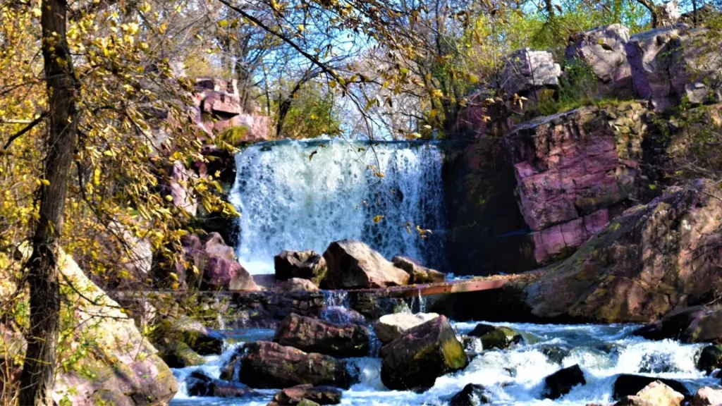 A photo of the Winnewissa Falls, a waterfall in Minnesota. It is surrounded by large boulders and craggy rocks, along with sparse foliage.